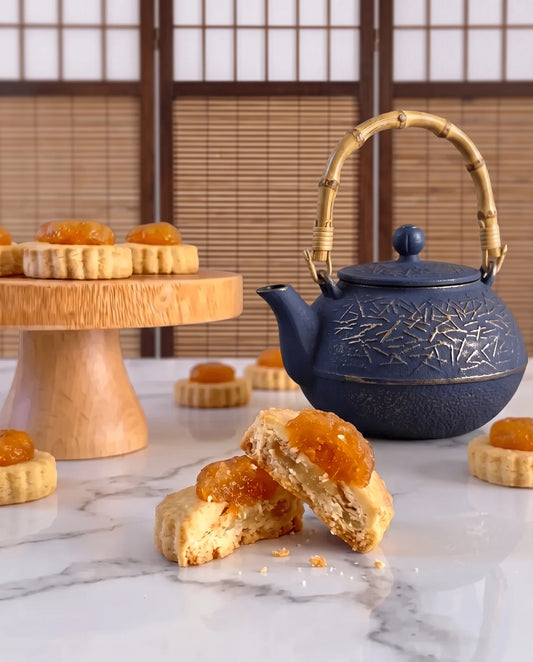 A dark blue cast iron teapot with a bamboo handle sits on a marble countertop next to golden pineapple tarts with more pineapple tarts on a wooden stand in the background.