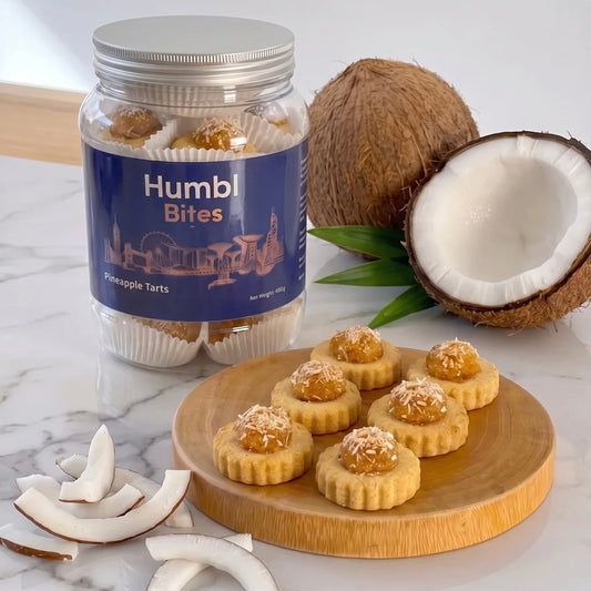 A jar of Humbl Bites Coconut Pineapple Tarts sits on a white marble surface next to whole and halved coconuts, with several tart pieces arranged on a round wooden board.