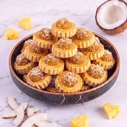 A stack of coconut pineapple tarts, presented in a dark bowl on a white marble surface. Slices of coconut and chunks of pineapple are arranged around it, with a halved coconut visible in the upper right.