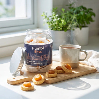 A jar of Humbl Bites pineapple tarts sits on a wooden board next to a steaming mug in a bright kitchen setting.