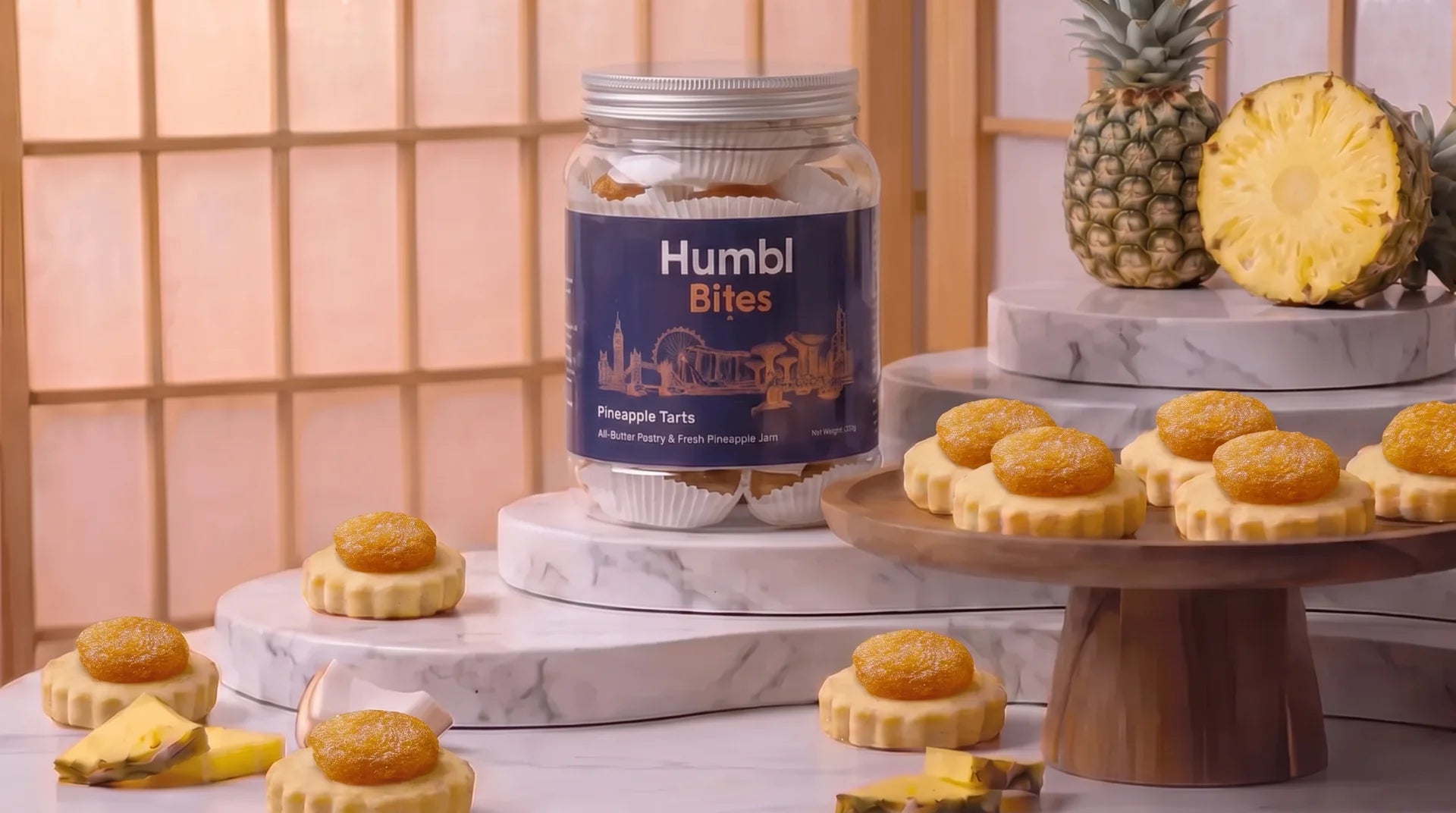A jar of Humbl Bites containing pineapple tarts, styled on marble risers with several baked pineapple tarts on stands and whole/half pineapples in the background.