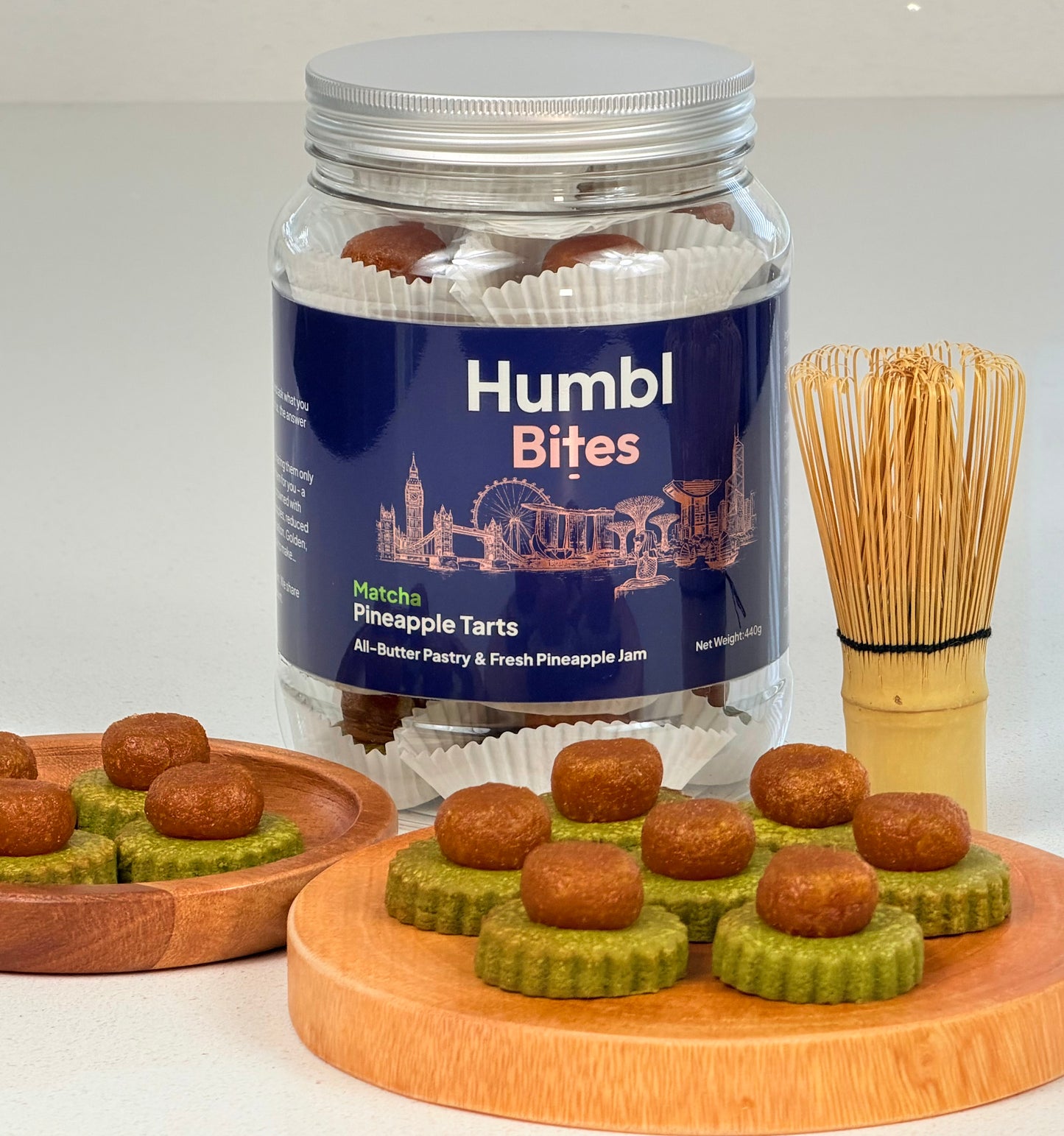 Jar of Humbl Bites pineapple tarts with a wooden tray and matcha whisk on a white background