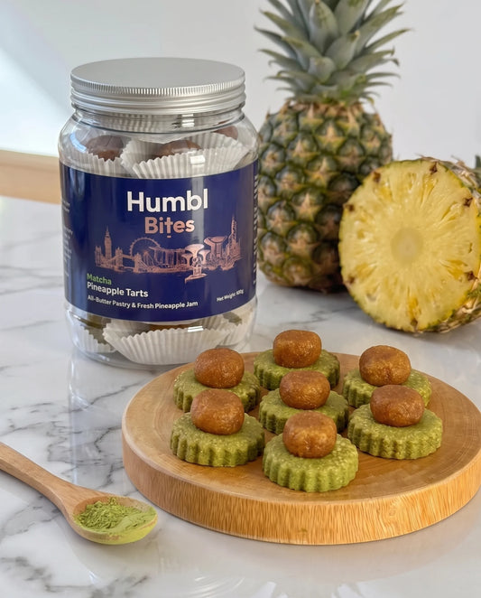 A jar of Humbl Bites pineapple tarts sits on a marble surface with several small pineapple tart pieces arranged on a round wooden board in front. Whole and halved pineapples are behind the jar, and a small spoonful of green powder is visible to the left.