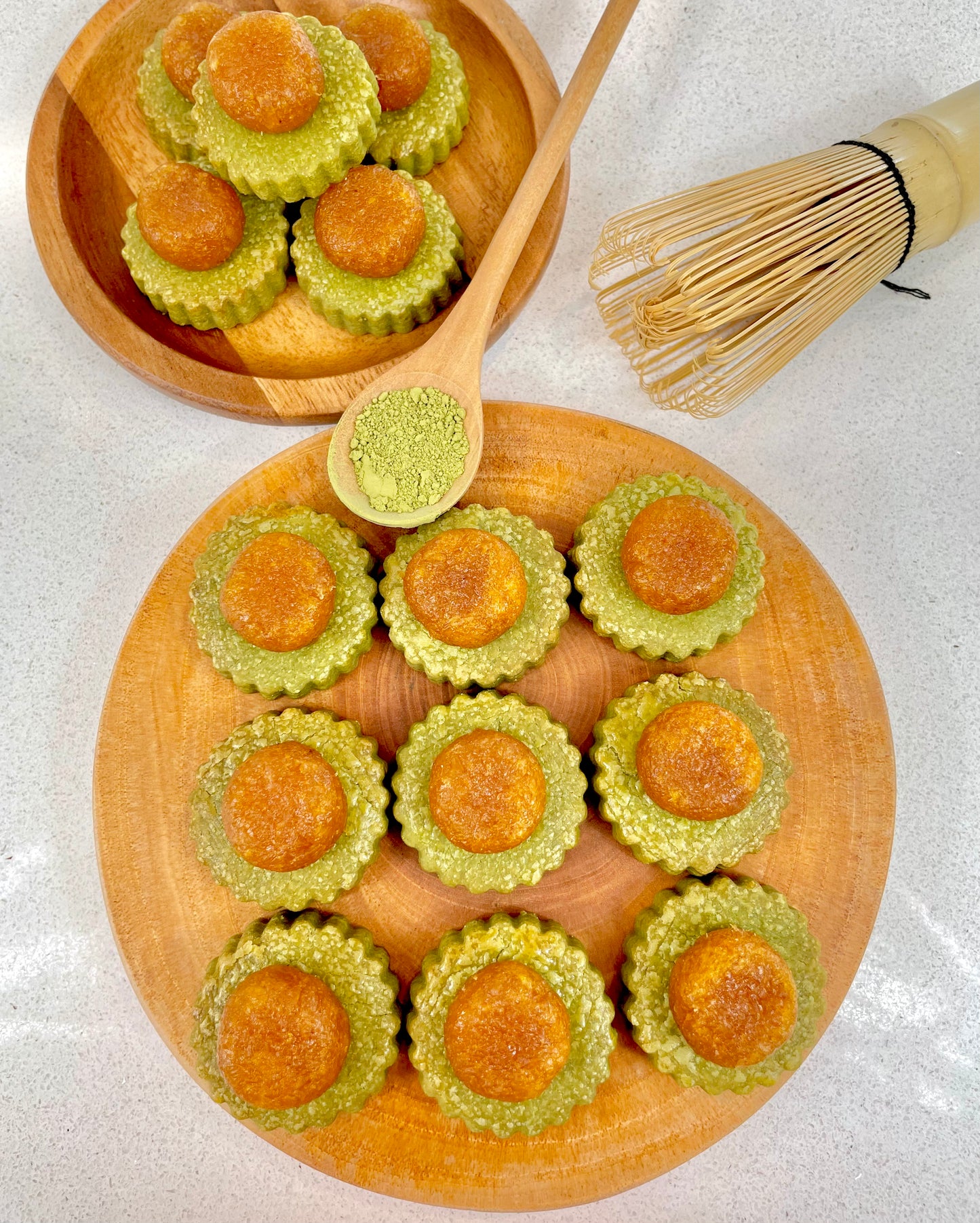 Wooden plate with matcha pineapple tarts, wooden spoon with green powder, and whisk on a light background