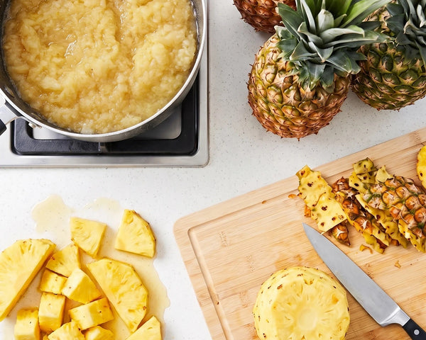 A kitchen scene showing pineapple being prepared: a pot of pineapple jam on a stove, whole pineapples, diced pineapple pieces, a cut pineapple on a wooden board, and a knife on a light countertop. New batch of jam for pineapple tarts