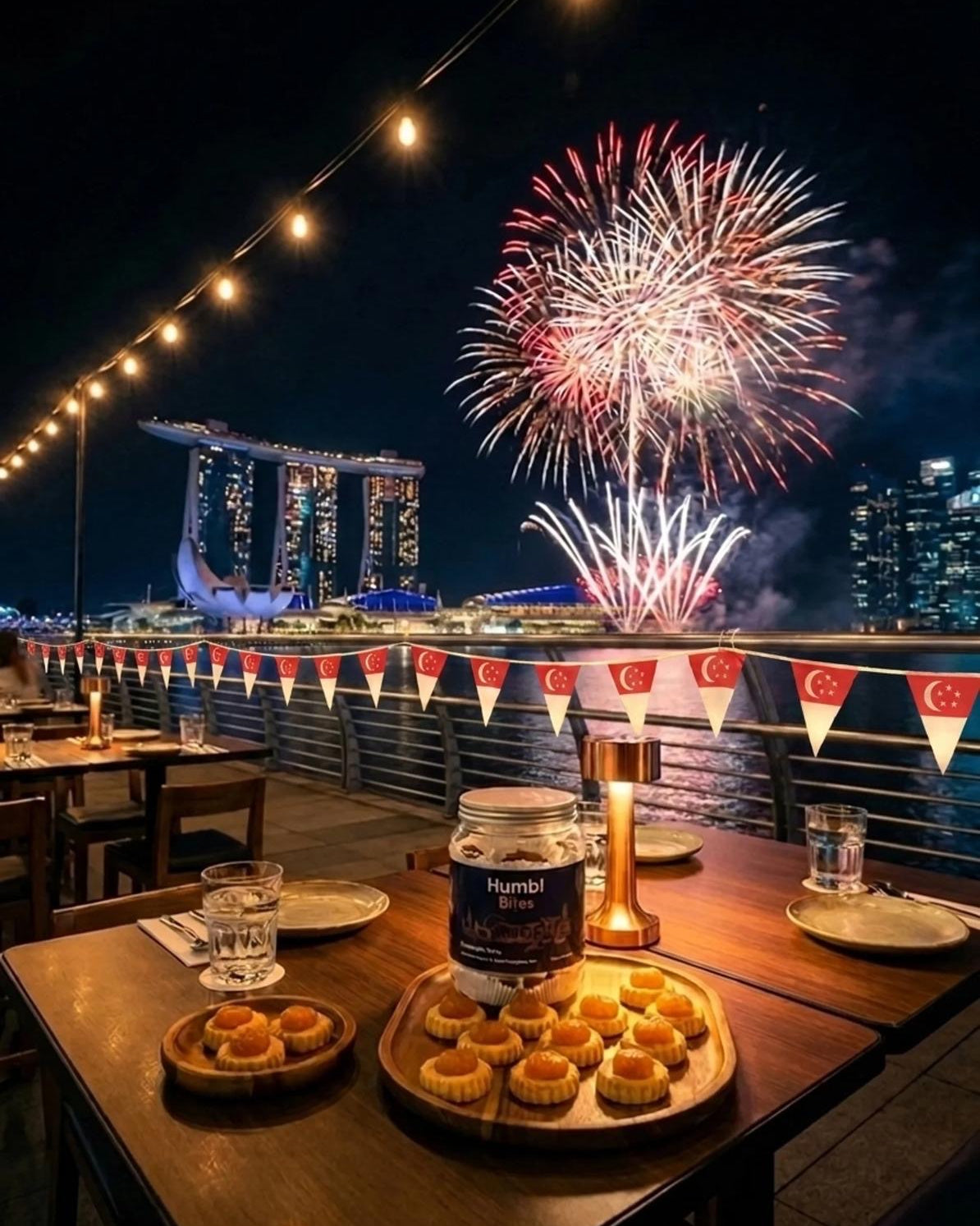 A jar labeled Humbl Bites with pineapple tarts placed on a wooden table in the foreground. In the foreground has a fireworks display