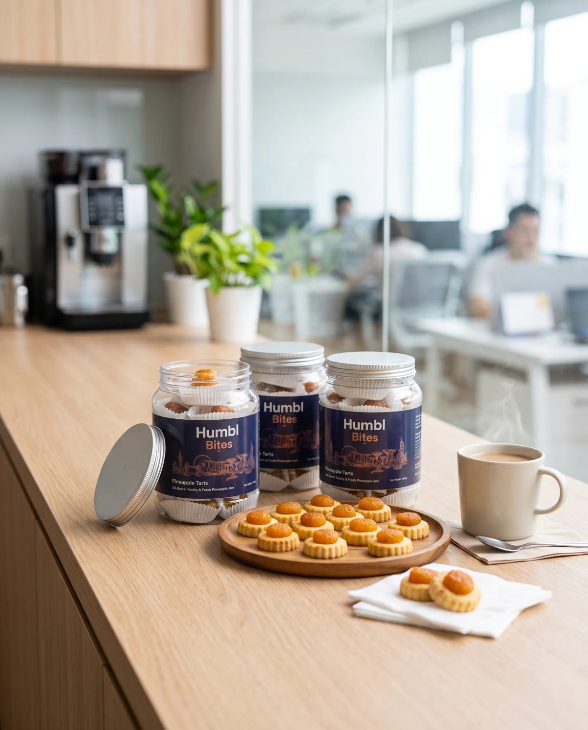 Three jars of Humbl Bites pineapple tarts arranged on a light wood counter in a modern office setting, alongside a coffee mug, and a few loose pastries on a napkin.
