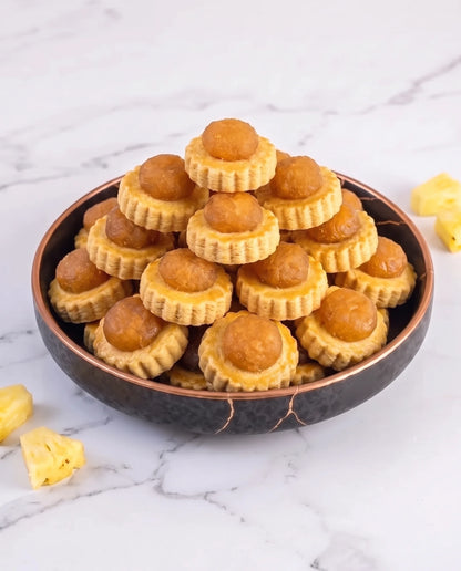 A stack of delicious pineapple tarts, presented in a dark ceramic bowl on a white marble surface with pineapple chunks scattered nearby.