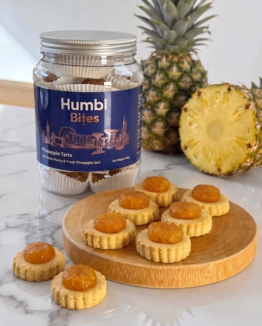 A jar of Humble Bites Pineapple Tarts stands on a white marble surface with several pineapple tart pieces arranged on a round wooden board and a whole pineapple plus a cut pineapple in the background.